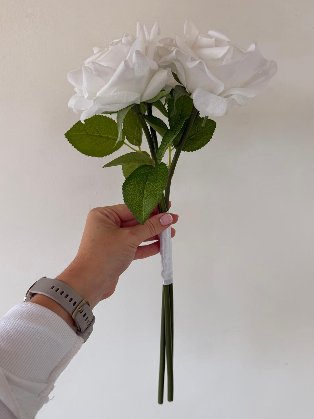 A hand holding a bouquet of white artificial roses with green leaves and stems against a plain off-white wall.