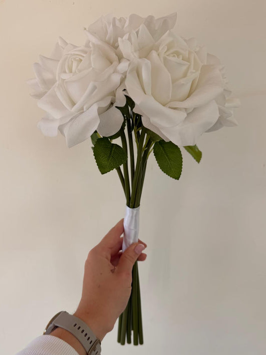 A hand holding a bouquet of three large white artificial roses with green stems and leaves against a plain off-white wall.